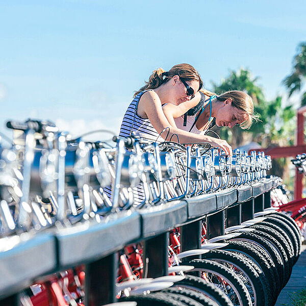 Zwei Personen beugen sich über eine lange Reihe ausgeliehener Fahrräder und prüfen die Räder an einer Station.