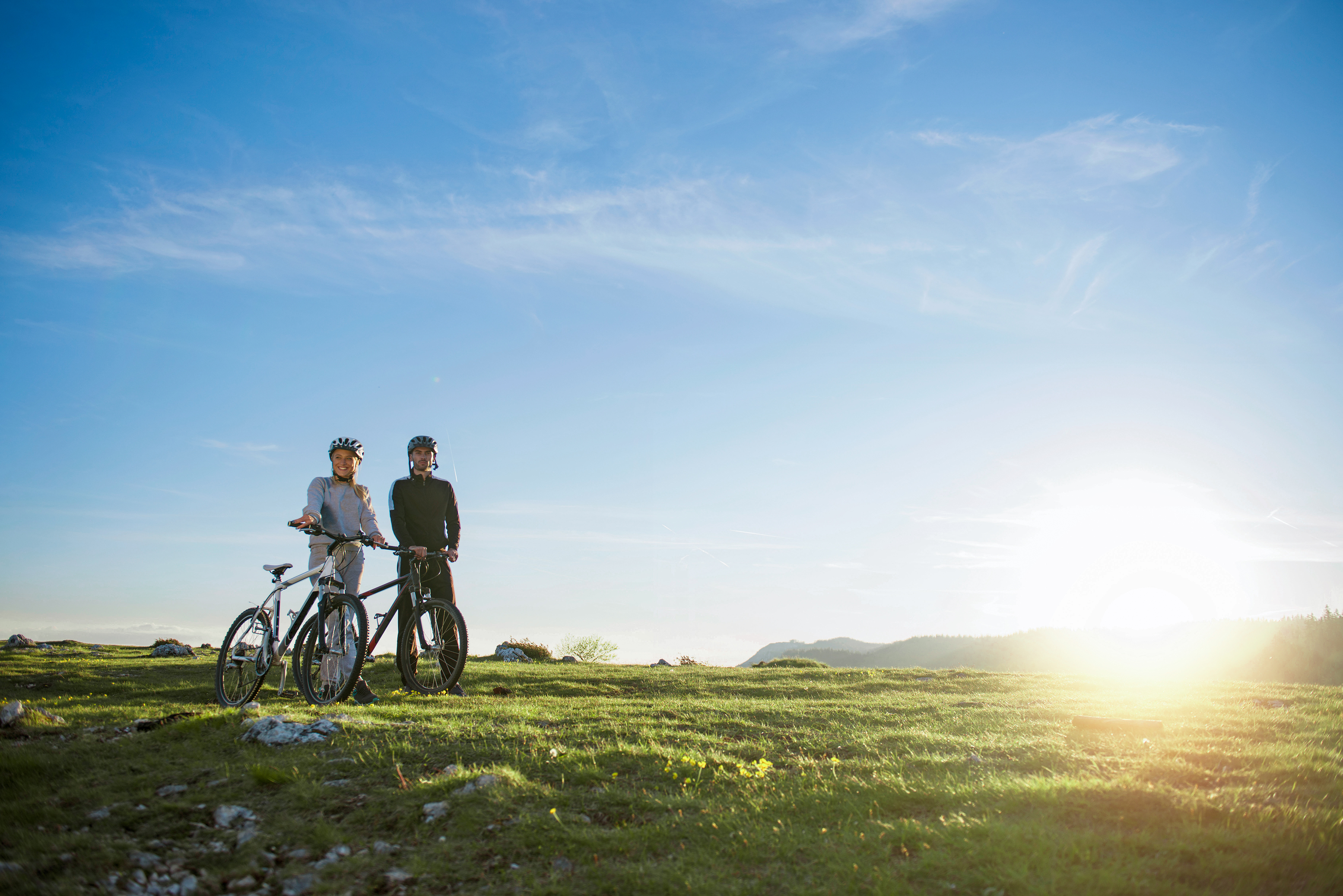 Zwei Menschen stehen mit ihren Fahrrädern auf einer Wiese. Sie blicken leicht nach links, während die Sonne tief am Himmel steht.