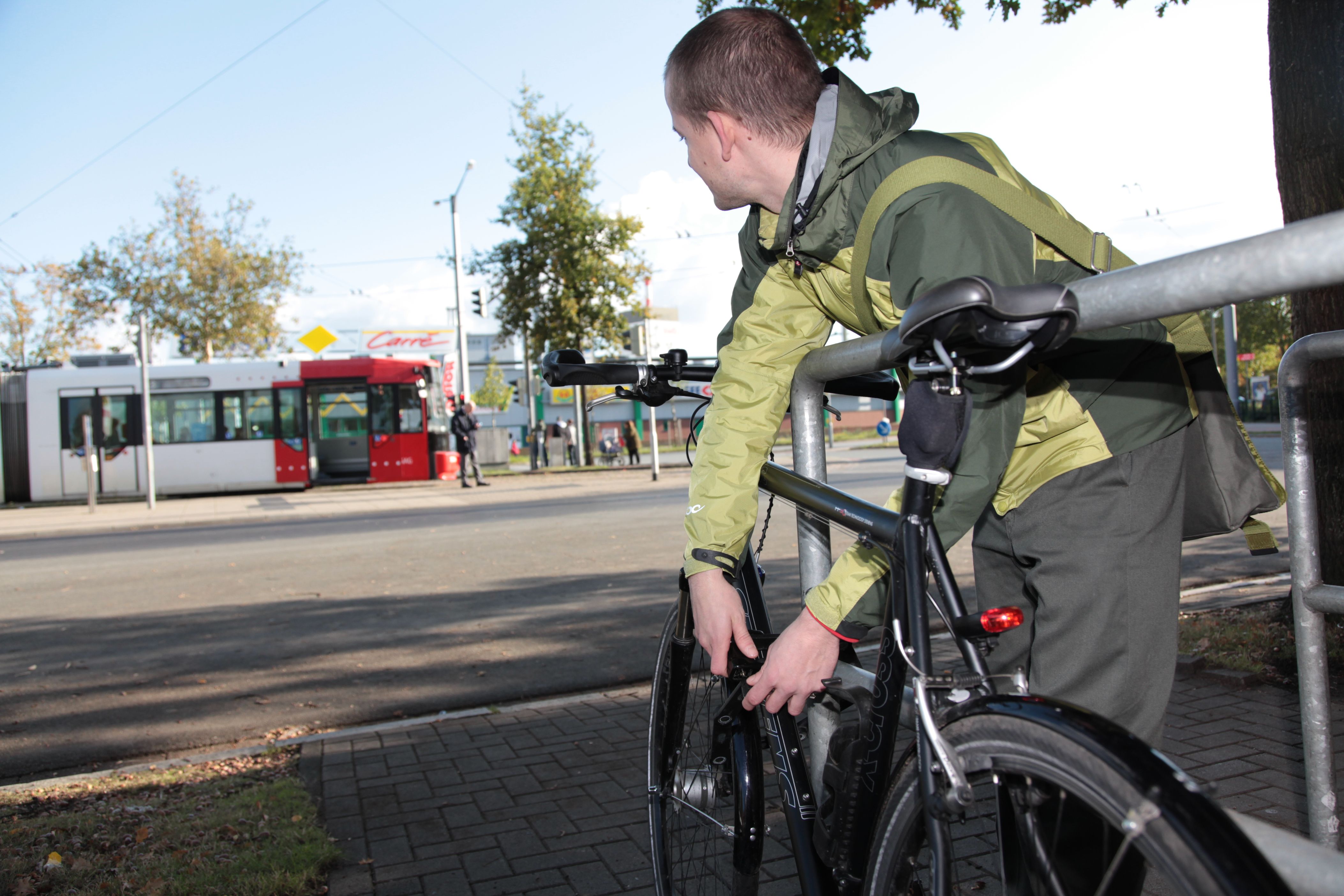 Ein junger mann schließt sein Fahrrad an einem Fahrradbügel an und schaut zu einer Straßenbahn im Hintergrund. 
