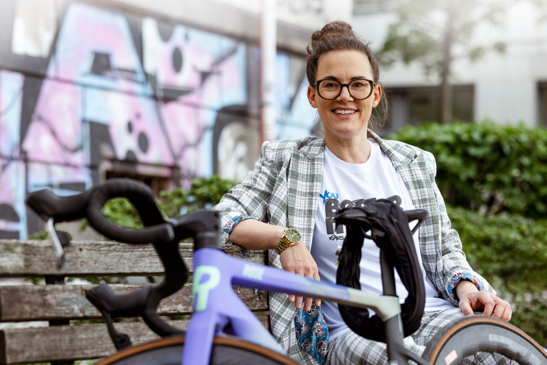 Eine Frau sitzt entspannt auf einer Holzbank neben einem violetten Rennrad vor einer Graffiti-Wand