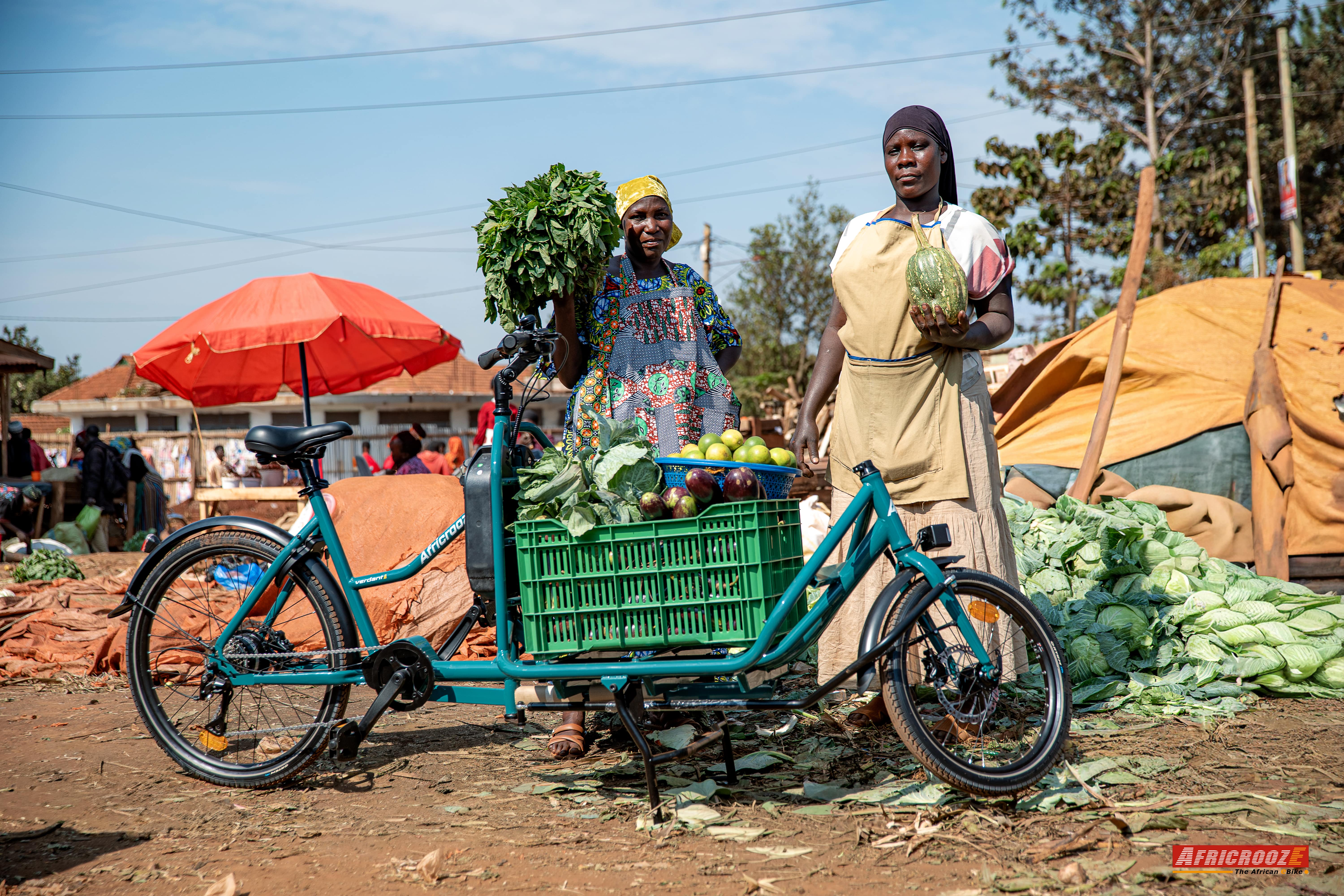 Zwei afrikanische Frauen stehen auf einem Markt im Freien hinter einem türkisgrünen Lastenfahrrad. Auf der tiefen Ladefläche zwischen Lenker und Vorderrad steht eine grüne Kiste, die voll mit Auberginen, Orangen und Blattgemüse beladen ist. Die Frauen halten weiteres Gemüse in den Händen und blicken freundlich in die Kamera.