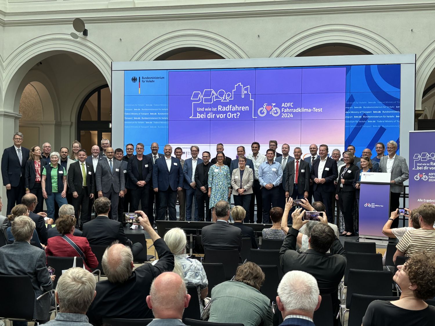 Auf einer großen Bühne im Bundesverkehrsministerium sind zahlreiche Städtevertreterinnen und -vertreter versammelt, eingerahmt von Bundesverkehrsminister Schnieder und ADFC-Bundesvorsitzenden Masurat. 