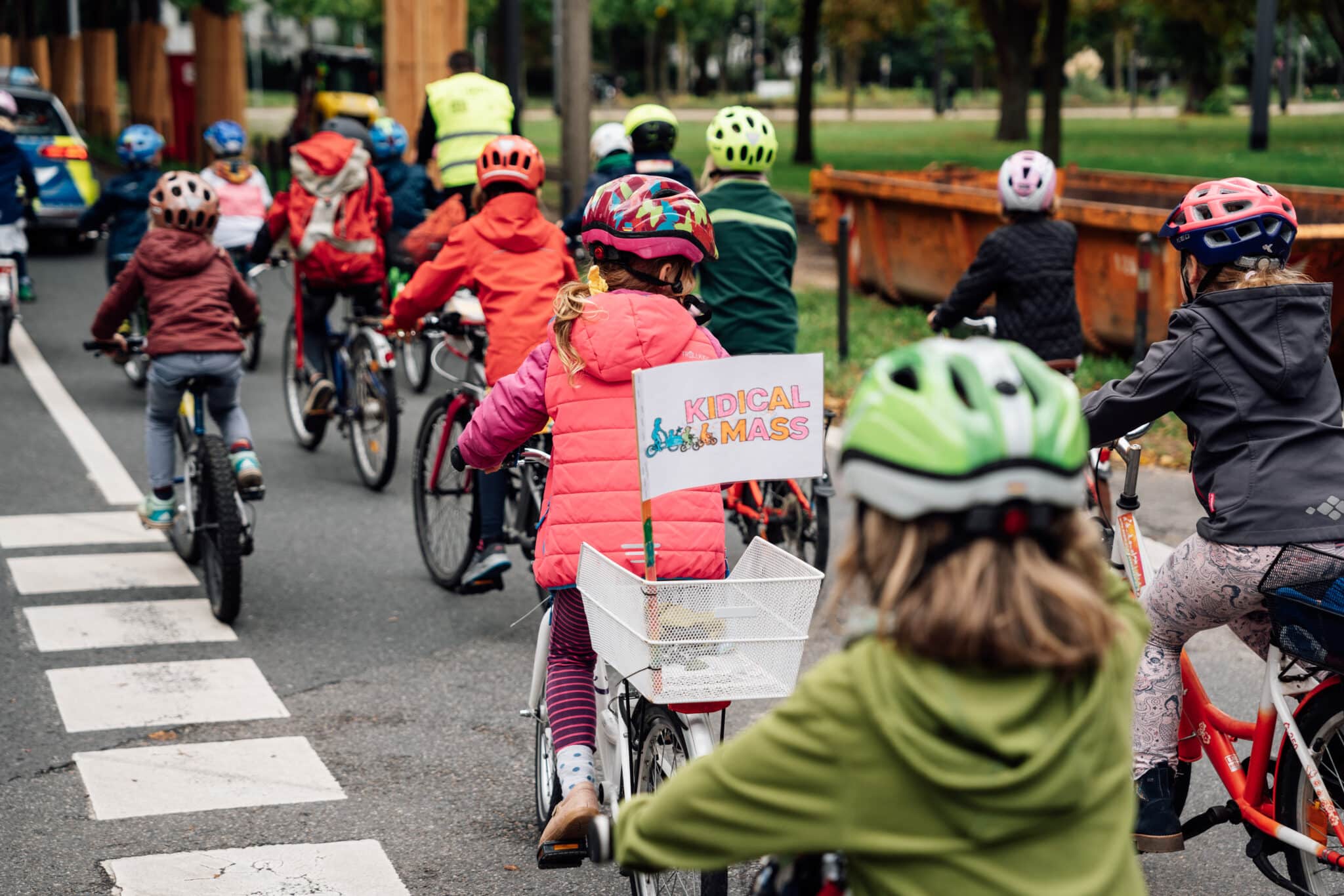 Mehrere Kinder und Erwachsene fahren bei einer Kidical Mass gemeinsam mit Fahrrädern und Helmen auf einer Straße. Ein Kind trägt eine Weste und hat ein Schild mit „Kidical Mass“ am Fahrradkorb.