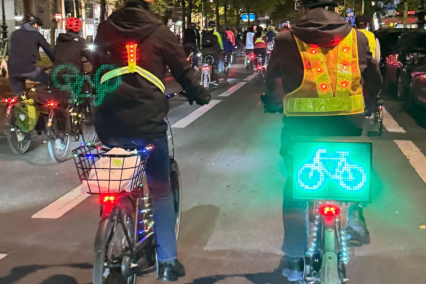 ADFC-Lichterfahrt Berlin, 10. Oktober 2025: Mehrere Menschen fahren auf beleuchteten Fahrrädern durch eine Fußgängerzone. An den Fahrrädern leuchten rote und grüne LED-Lichter, ein Fahrrad trägt ein leuchtend grünes Display mit Fahrrad-Symbol. Die Radfahrenden tragen fluoreszierende Sicherheitswesten. Im Hintergrund sind weitere beleuchtete Fahrräder zu sehen.