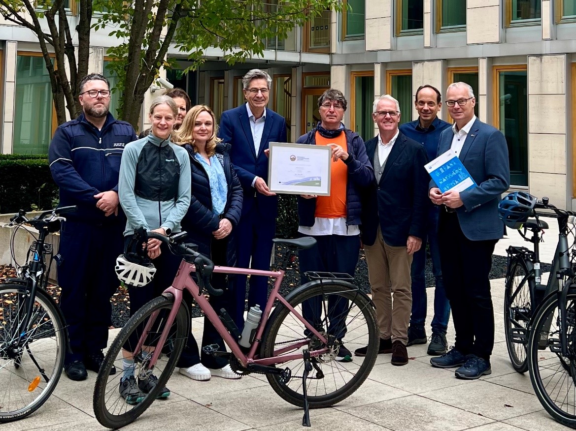 Gruppe von neun Personen steht vor dem Oberlandesgericht Düsseldorf, in der Mitte wird das gerahmte Gold-Zertifikat „Fahrradfreundlicher Arbeitgeber" präsentiert, im Vordergrund stehen drei Fahrräder.