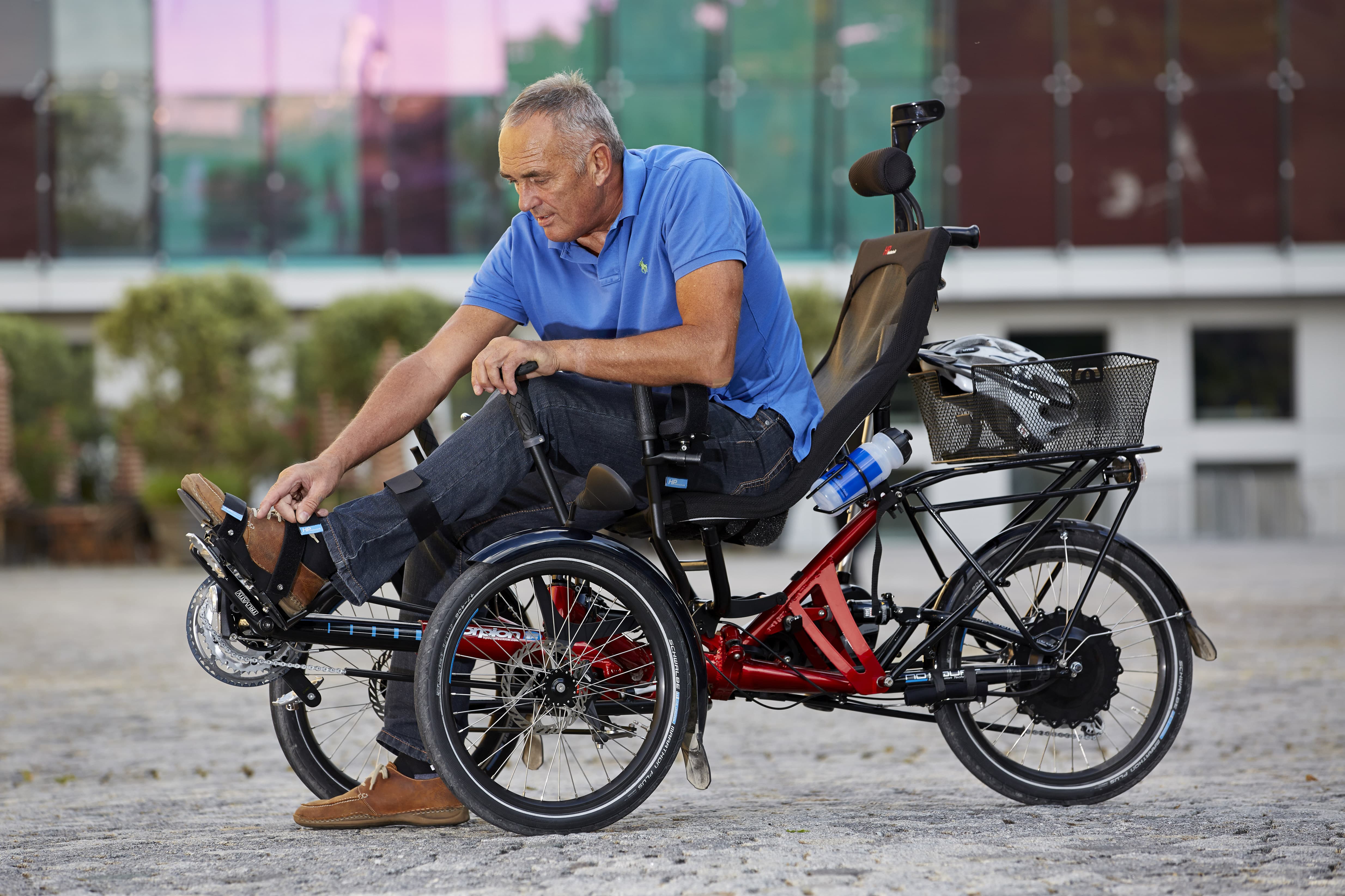 Ein Mann in blauem Poloshirt sitzt auf einem roten dreirädrigen Elektrofahrrad mit schwarzer Rückenlehne und Korb. Das Spezialrad steht auf einem gepflasterten Platz vor einem modernen Gebäude mit grünen Fenstern.