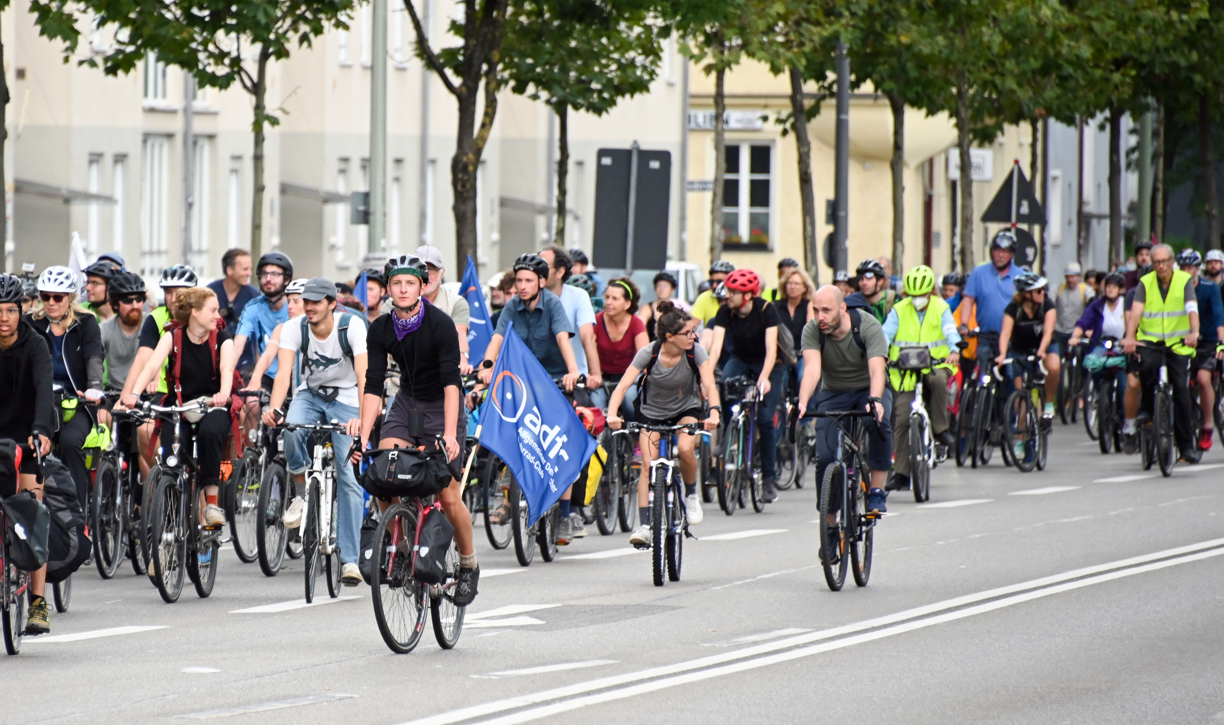 Teilnehmende der ADFC-Fahrradsternfahrt mit blauer ADFC-Fahne fahren gemeinsam auf einer breiten Straße. Menschen jeden Alters mit Helmen und teilweise Warnwesten demonstrieren für bessere Radinfrastruktur. Im Hintergrund säumen Bäume die Straße.
