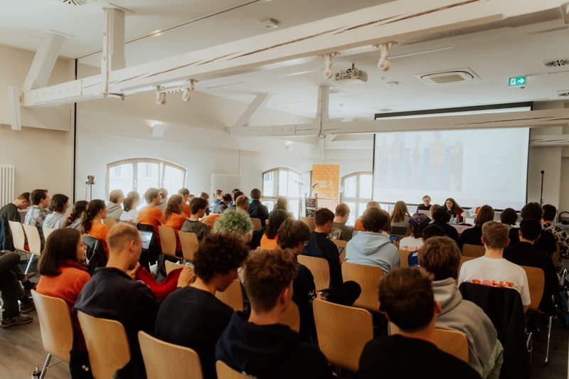 Junge Menschen sitzen auf Stühlen in einem große Saal und schauen nach vorn auf eine Leinwand.