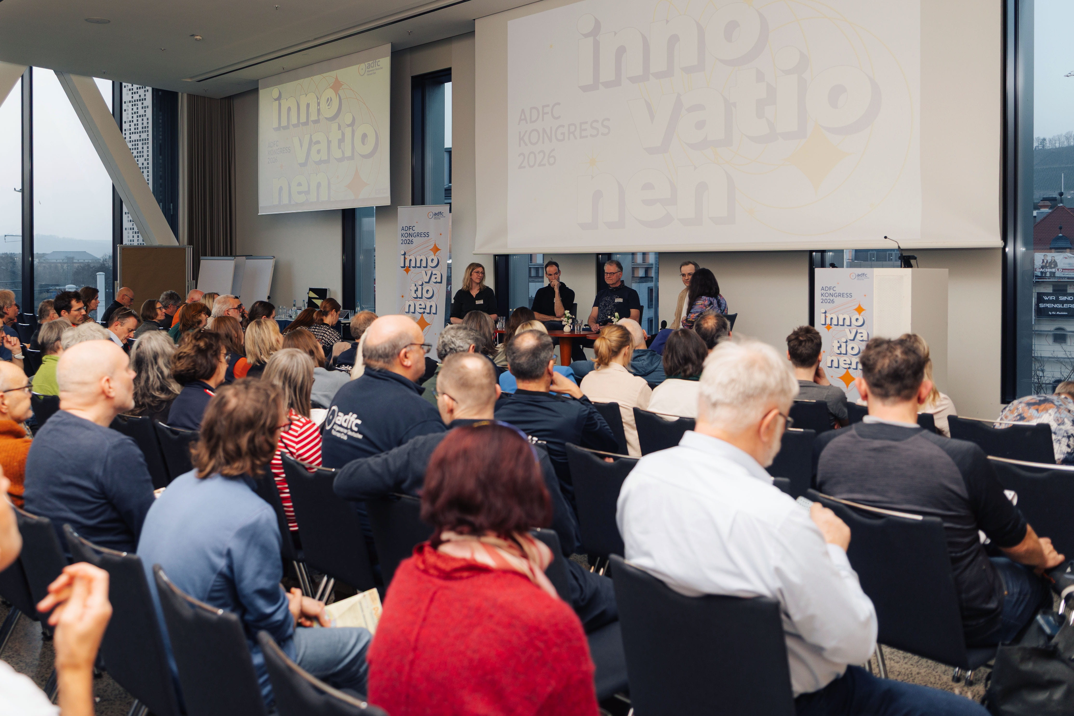 Blick über die Köpfe zahlreicher sitzender Zuschauer im Saal auf eine Bühne. Dort diskutieren vier Personen bei einer Podiumsdiskussion. Im Hintergrund zeigen Leinwände den Schriftzug ADFC Kongress 2026 Innovationen.