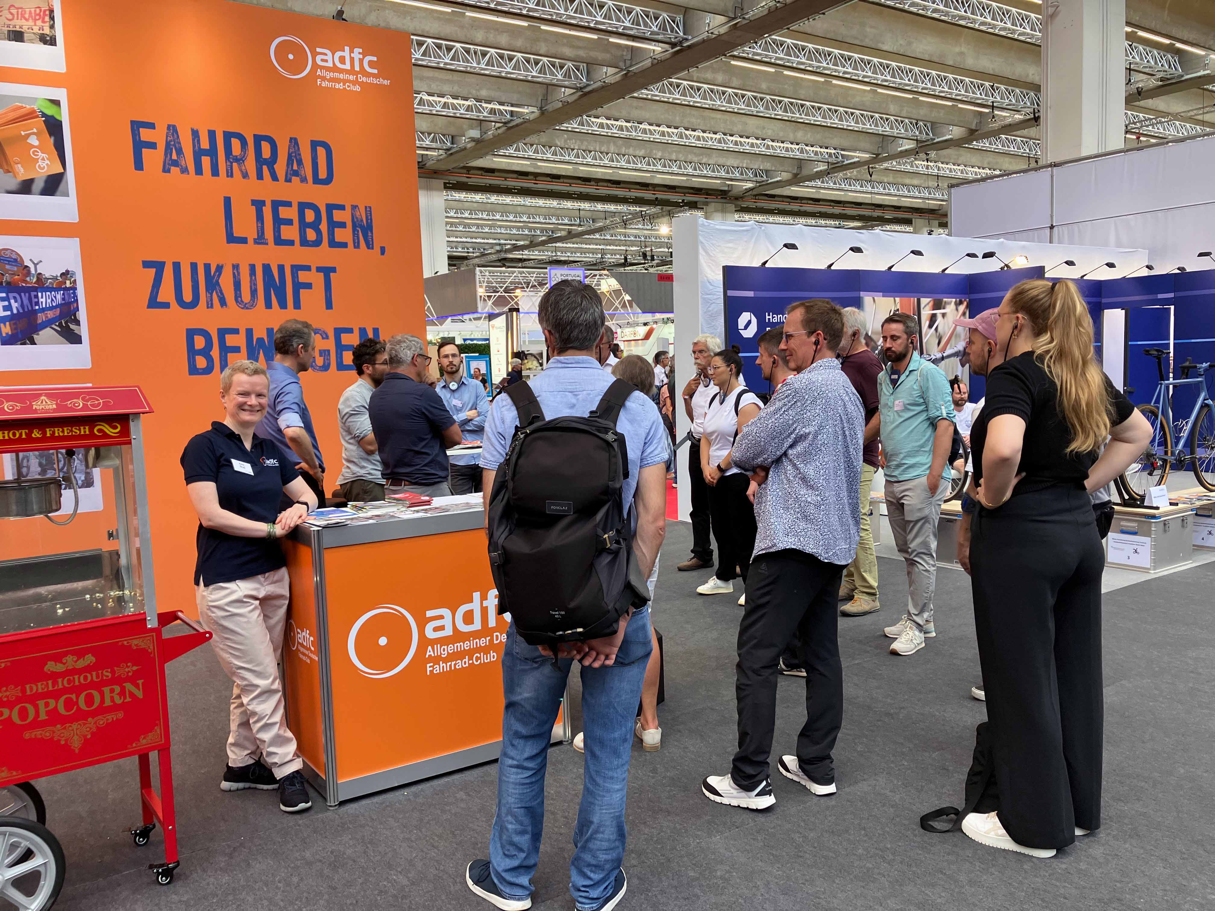Ein Messestand des ADFC in einer großen Halle. Vor einer orangen Rückwand mit dem blauen Slogan „Fahrrad lieben, Zukunft bewegen“ steht eine lächelnde Frau im dunkelblauen ADFC-Shirt an einer Theke. Mehrere Menschen stehen davor und dahinter in Gruppen zusammen und unterhalten sich. Im Vordergrund links ist eine rote Popcornmaschine zu sehen.