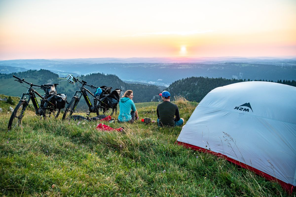 Zwei Radreisende sitzen in der Dämmerung neben einem Zelt im Grünen und blicken auf einen Höhenzug in der Ferne. 
