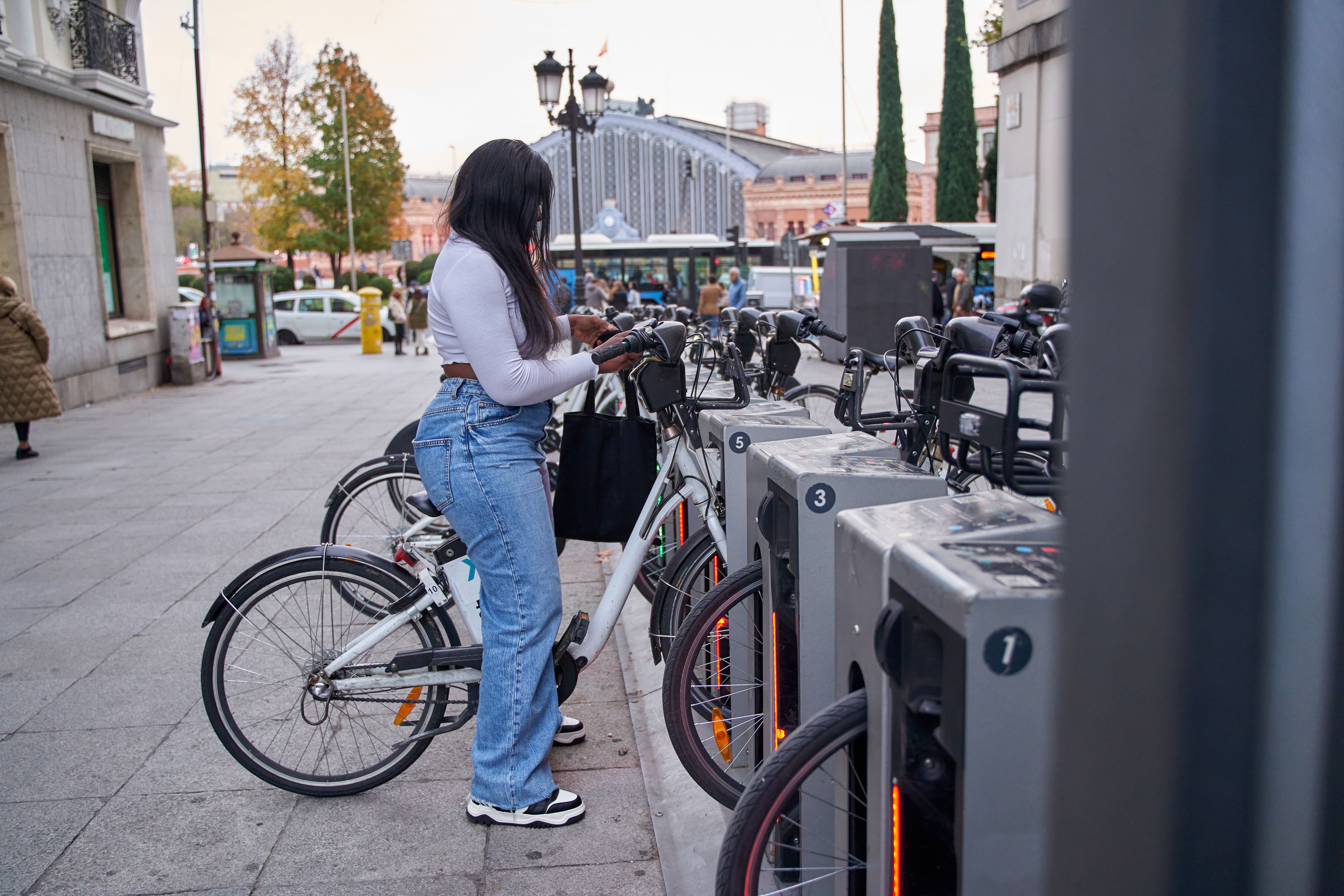 Eine junge Frau mit langen dunklen Haaren, weißem Oberteil, Jeans und schwarzer Umhängetasche steht an einer Bike-Sharing-Station und entnimmt ein weißes Leihfahrrad. Die Station verfügt über mehrere nummerierte Stellplätze mit grauen Dockingelementen. Im Hintergrund sind ein belebter Stadtplatz, historische Gebäude, Passanten und ein Bahnhofsgebäude zu sehen.