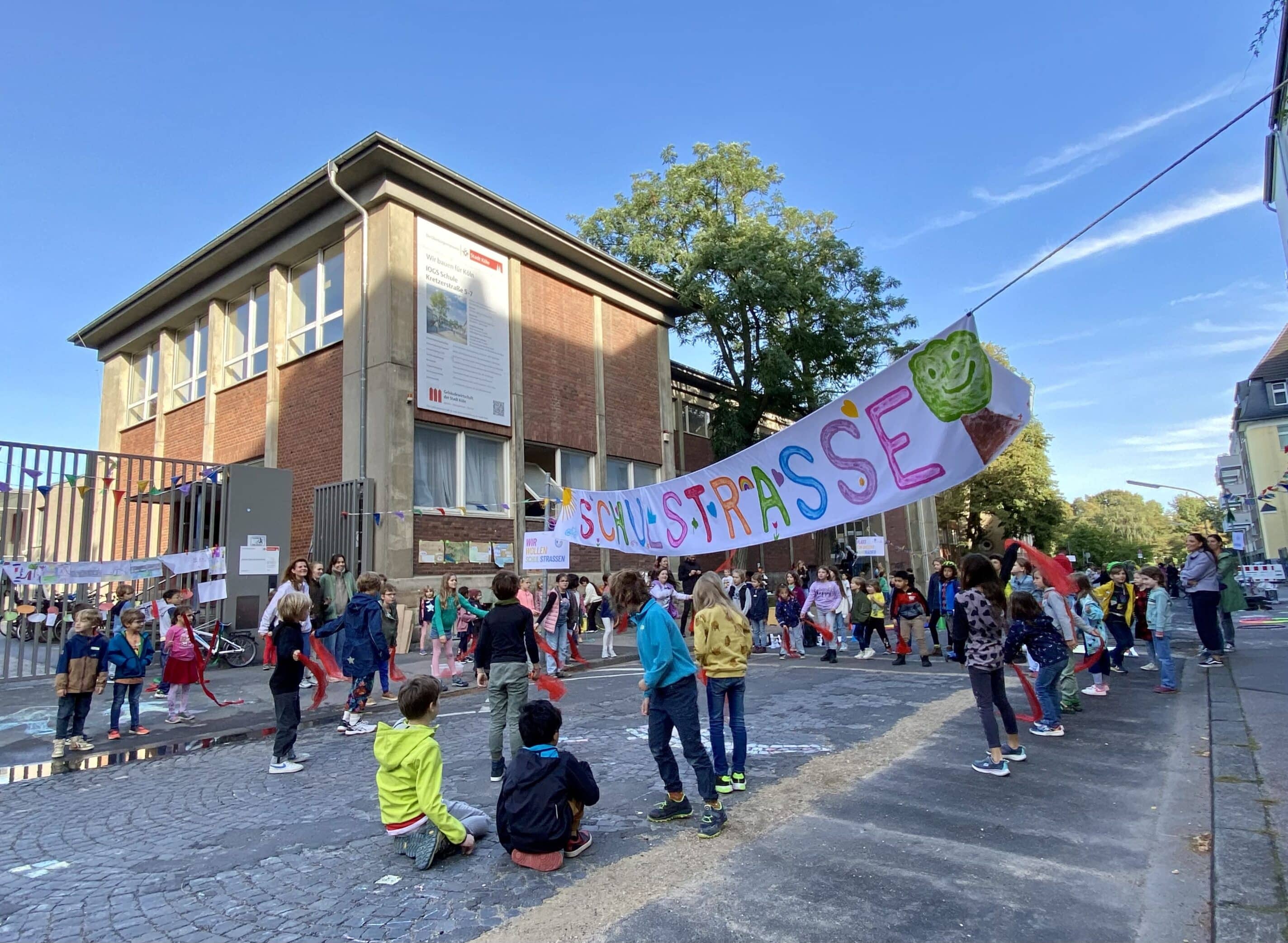 Kinder verschiedenen Alters spielen und bewegen sich auf einer gepflasterten Straße vor einem zweistöckigen Backsteingebäude. Über der Straße hängt ein buntes Banner mit der Aufschrift „Schulstraße“ und einem grünen Baum-Symbol. Im Hintergrund sind Erwachsene zu sehen, die die Aktion begleiten.