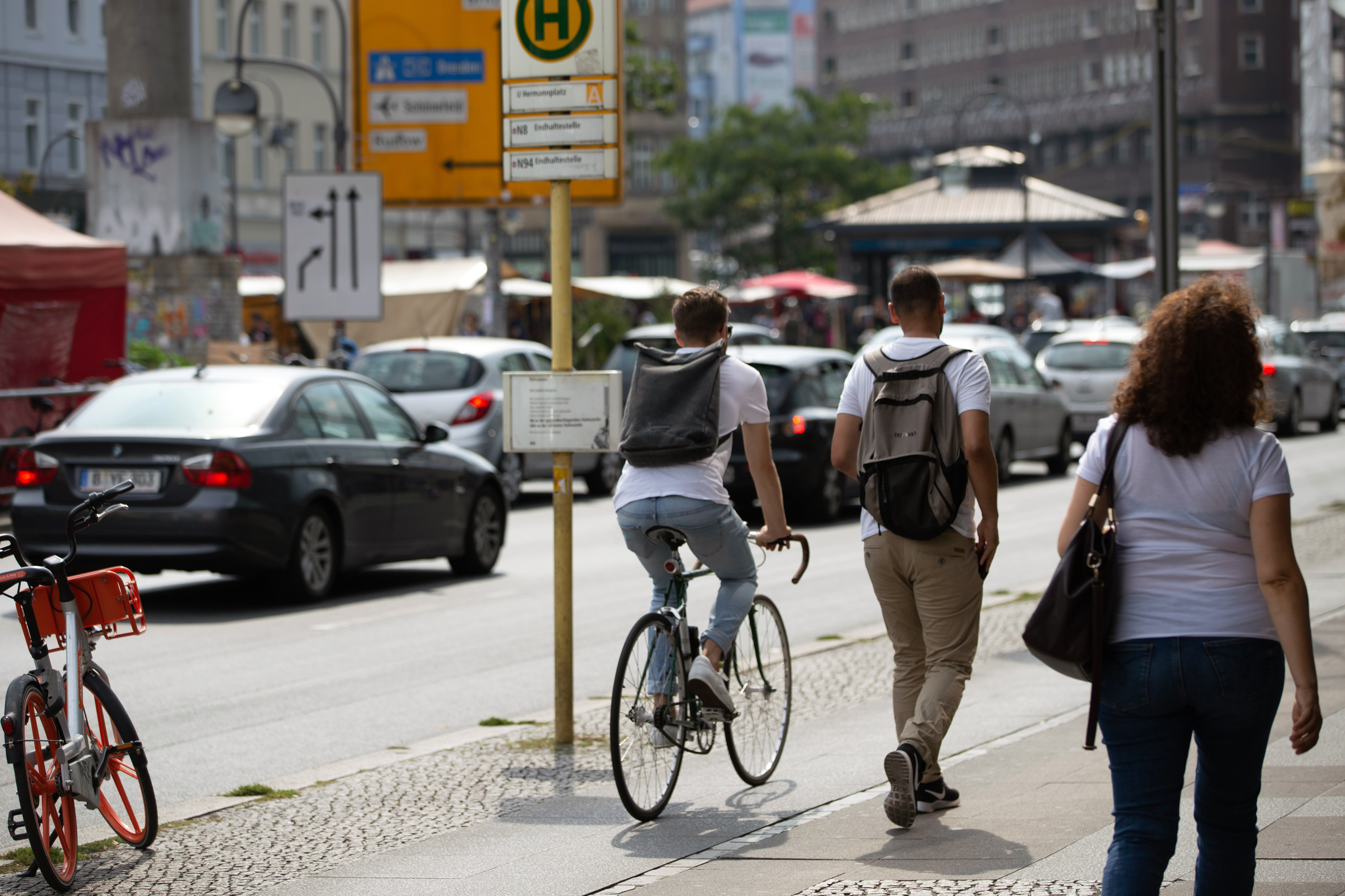 Eine Straßenszene: Ein Fahrradfahrer und zwei Fußgänger teilen sich einen gepflasterten Gehweg. Im Hintergrund parken Autos am Straßenrand.