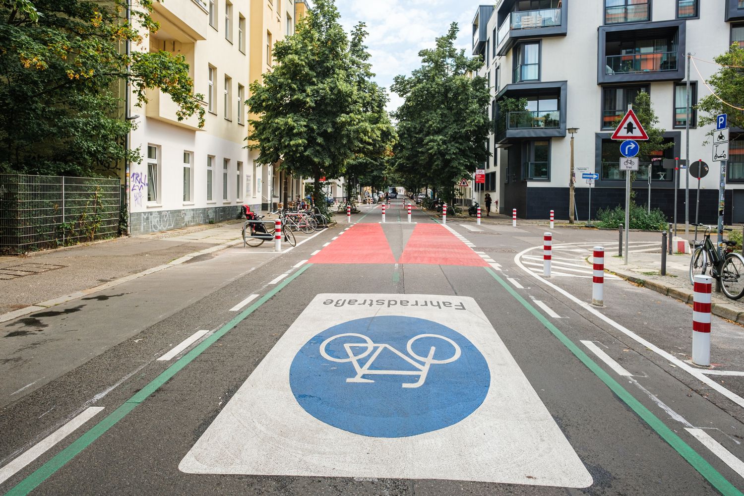 Eine Straße in Berlin: Rechts und links sind Häuserzeilen zu sehen, die Fahrbahn ist mit einem großen Fahrradsymbol versehen. Weiter hinten ist die Fahrbahn rot eingefärbt, Poller dienen als modaler Filter.  