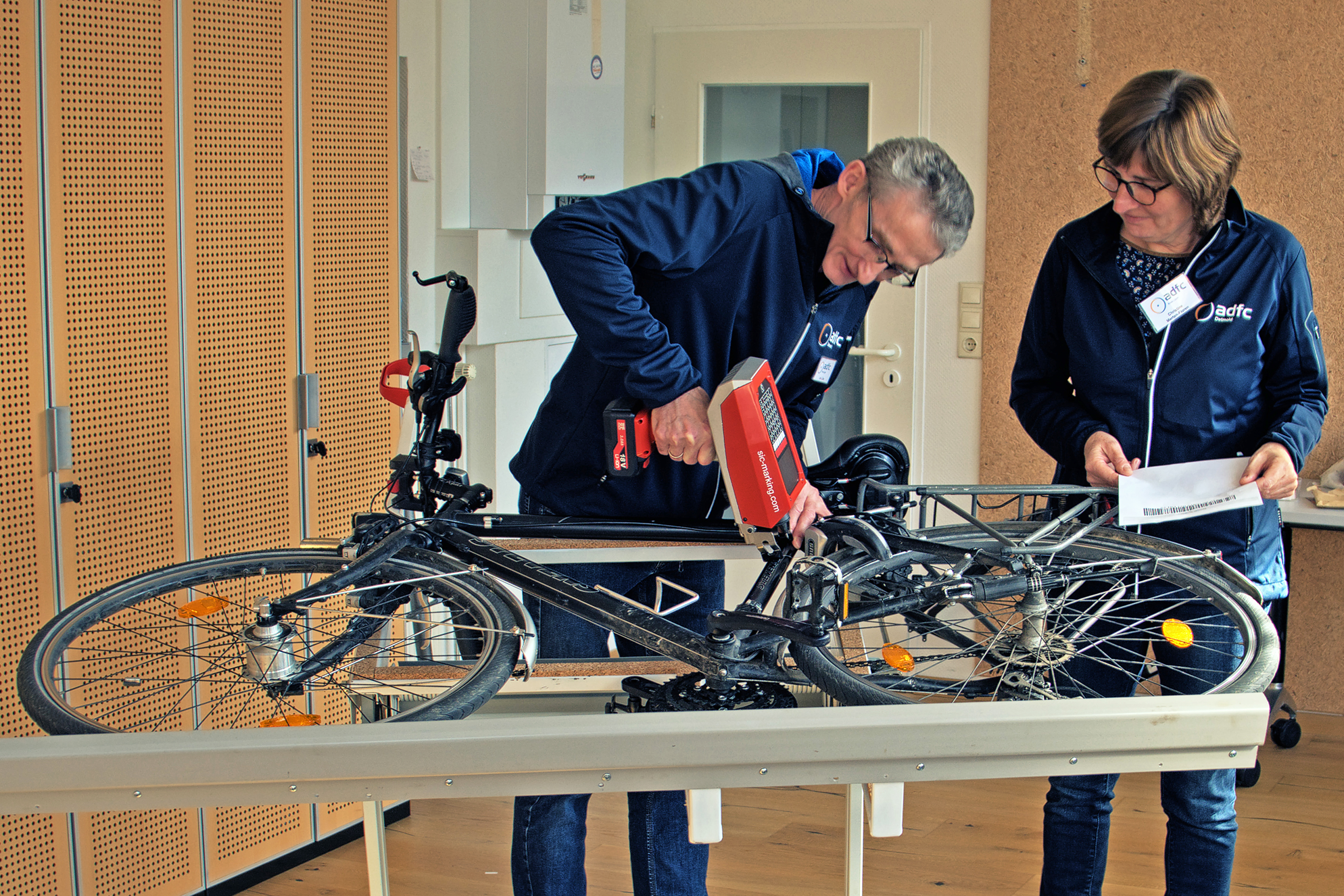 Ein Mann codiert mit einem Nadeldrucker ein auf der Seite liegendes Fahrrad, während die Besitzerin zuschaut. 