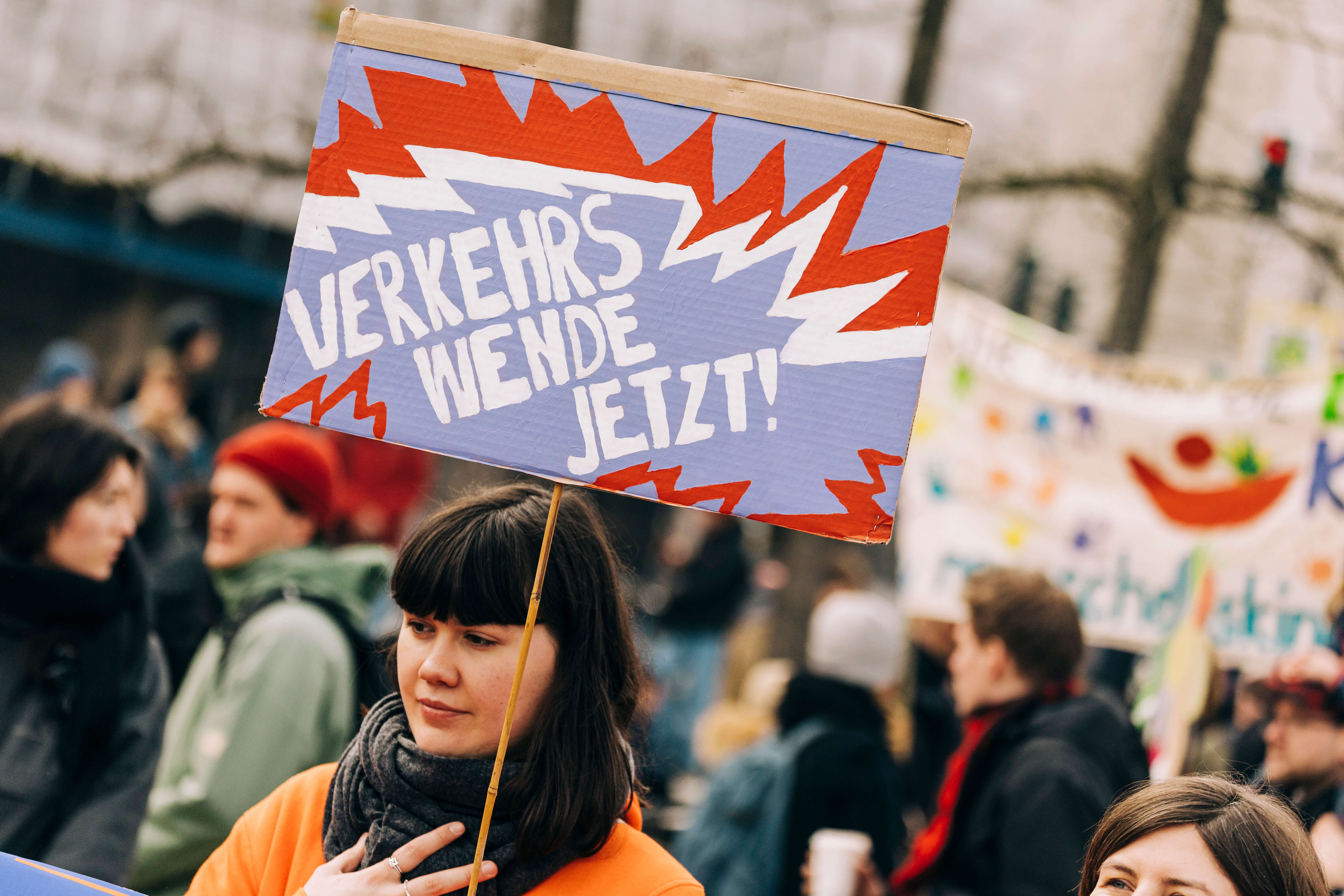 Junge Frau auf einer Demo hält ein Schild in die Luft, auf dem steht "Verkehrswende Jetzt!" 