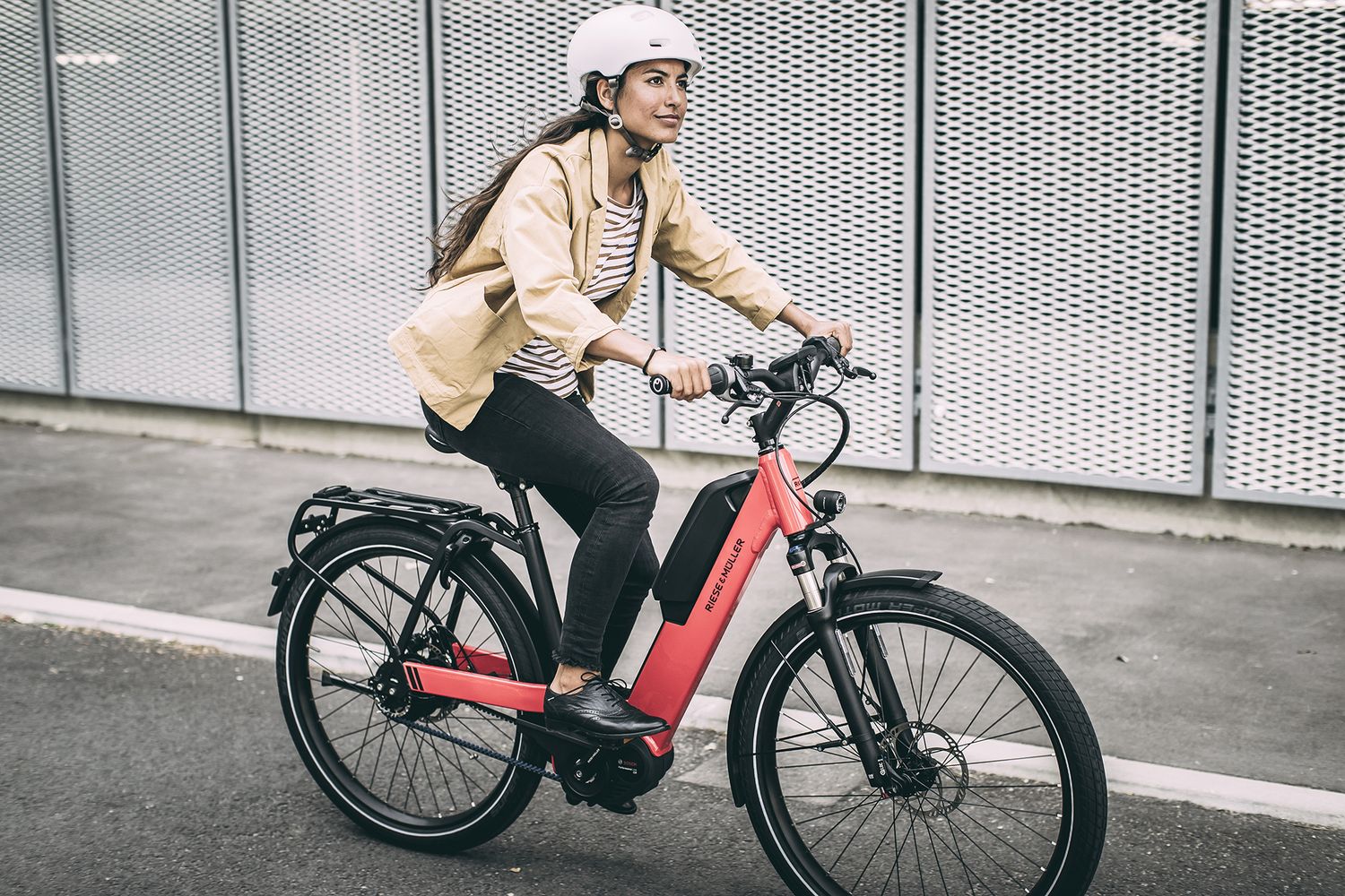 Eine Frau fährt auf einem roten Elektrorad auf Asphalt, im Hintergrund graue, gelochte Metallwände. 