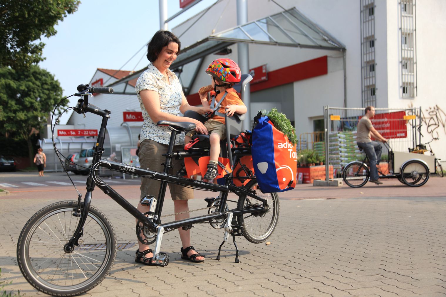 Frau schnallt Kind im Kindersitz an. Es sitzt hinten auf dem Fahrrad, dahinter sind noch Gepäcktaschen angehängt.