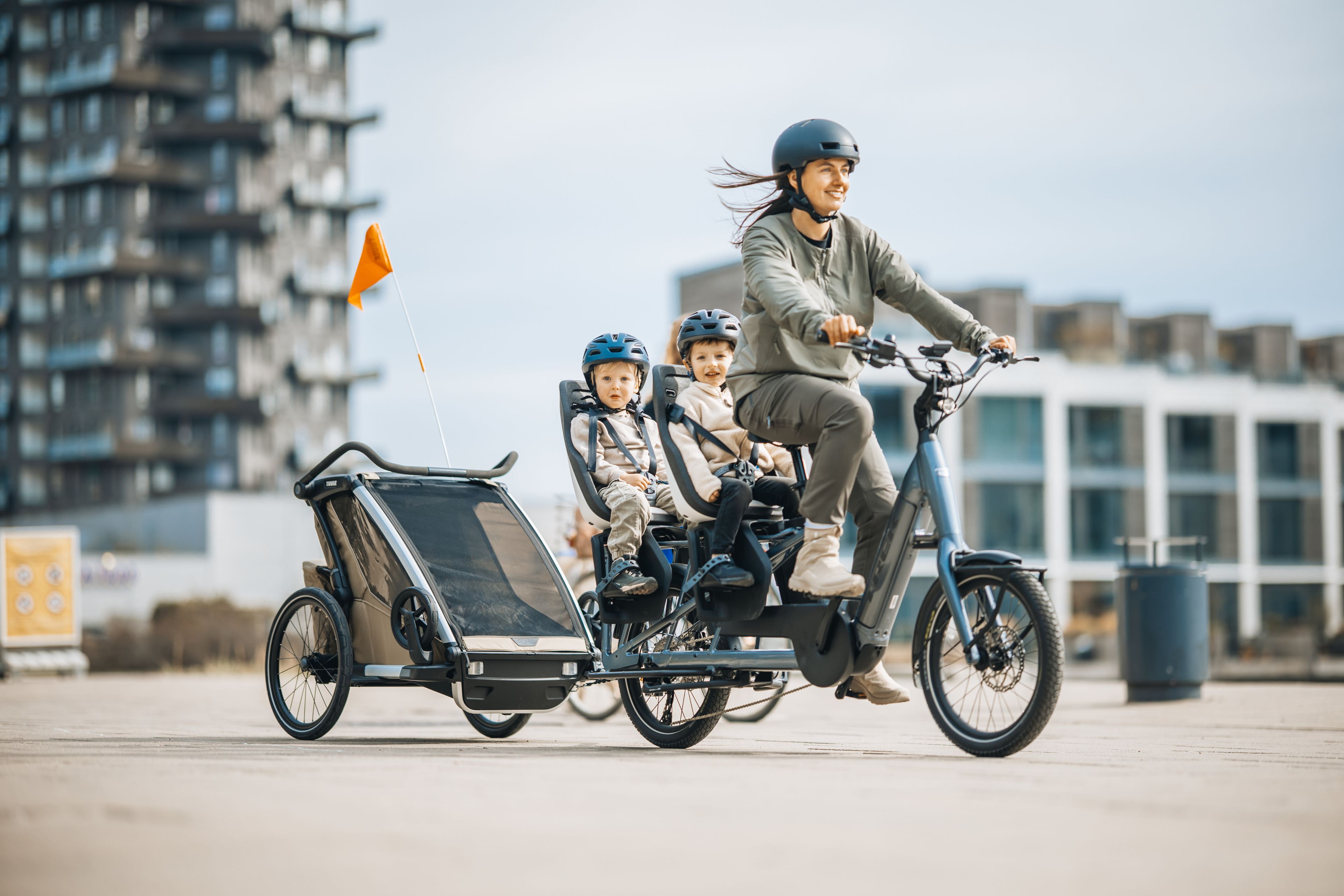 Frau fährt ein Lastenrad Longtail, auf dem langen Gepäckträger hinten sitzen zwei Kinder in je einem Kindersitz. Das Fahrrad zieht zudem einen Kinderfahrradanhänger.