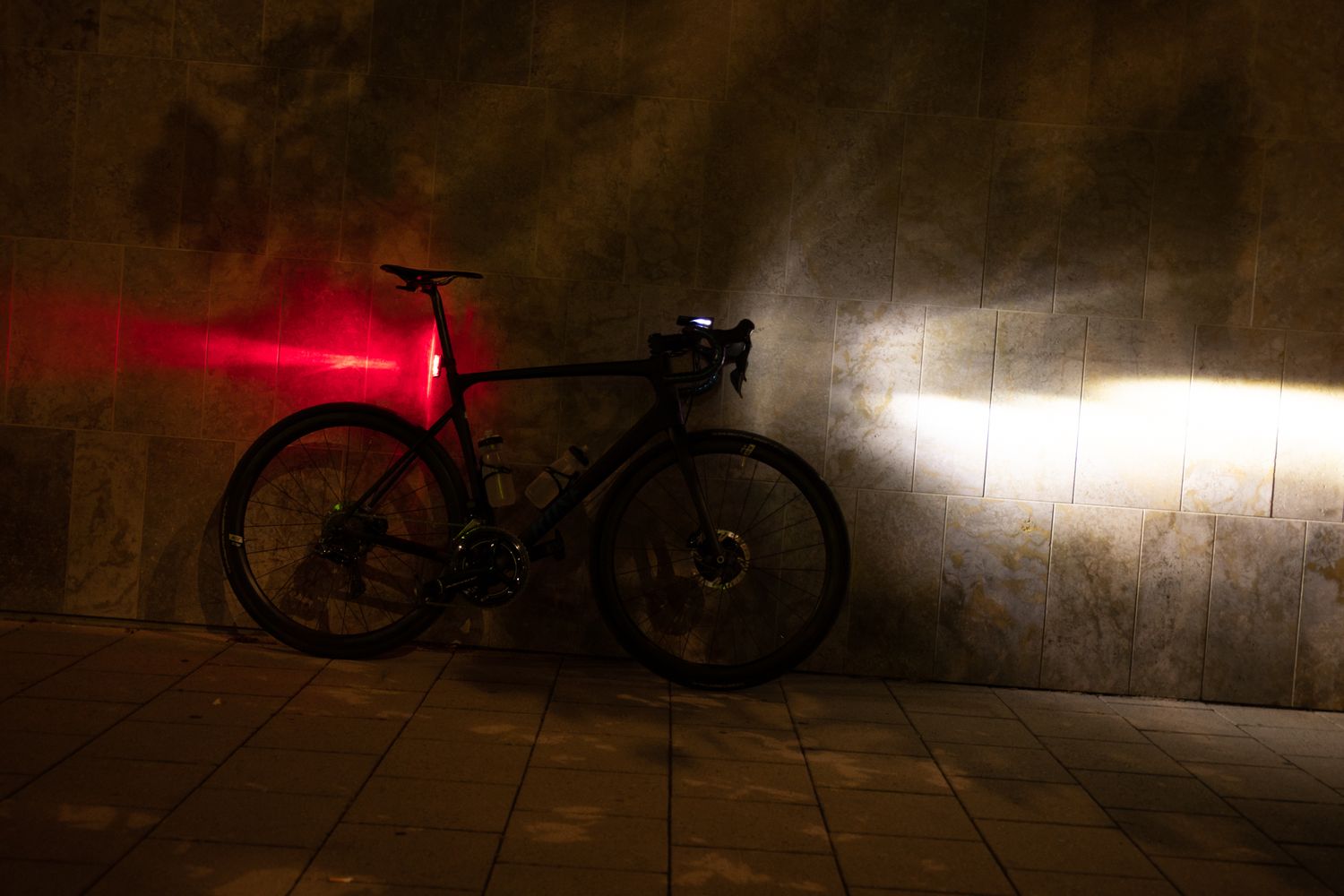Ein Fahrrad steht in der Dunkelheit an einer Wand, mit am Lenker montiertem, eingeschalteten Scheinwerfer und an der Sattelstütze montierten, leuchtenden Rücklicht.