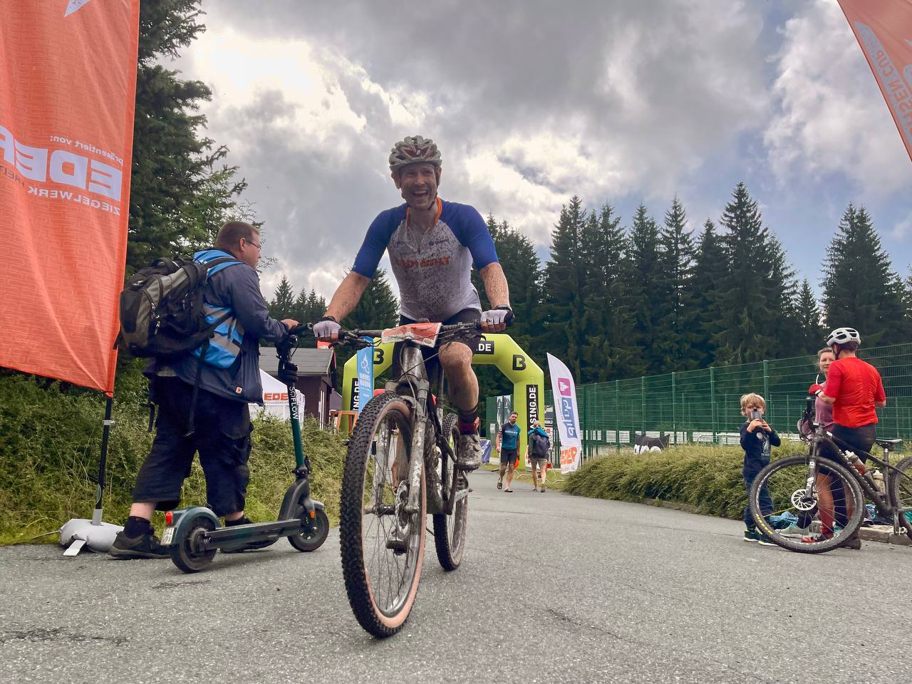 Ein mit schlamm bespritzter Mountainbiker hat gerade einen Zielbogen passiert und lächelt. 