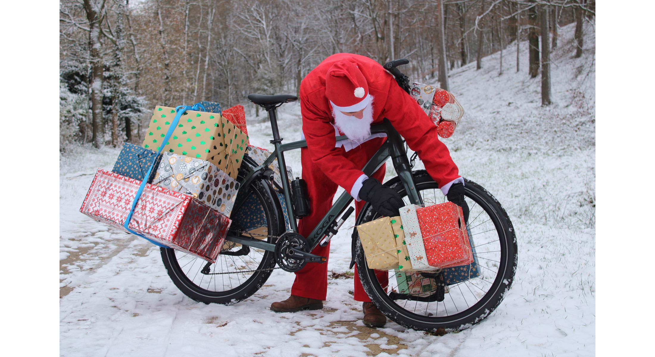 Man in Weihnachtskleidung beugt sich über ein mit Geschenken beladenes Fahrrad im Schnee. 