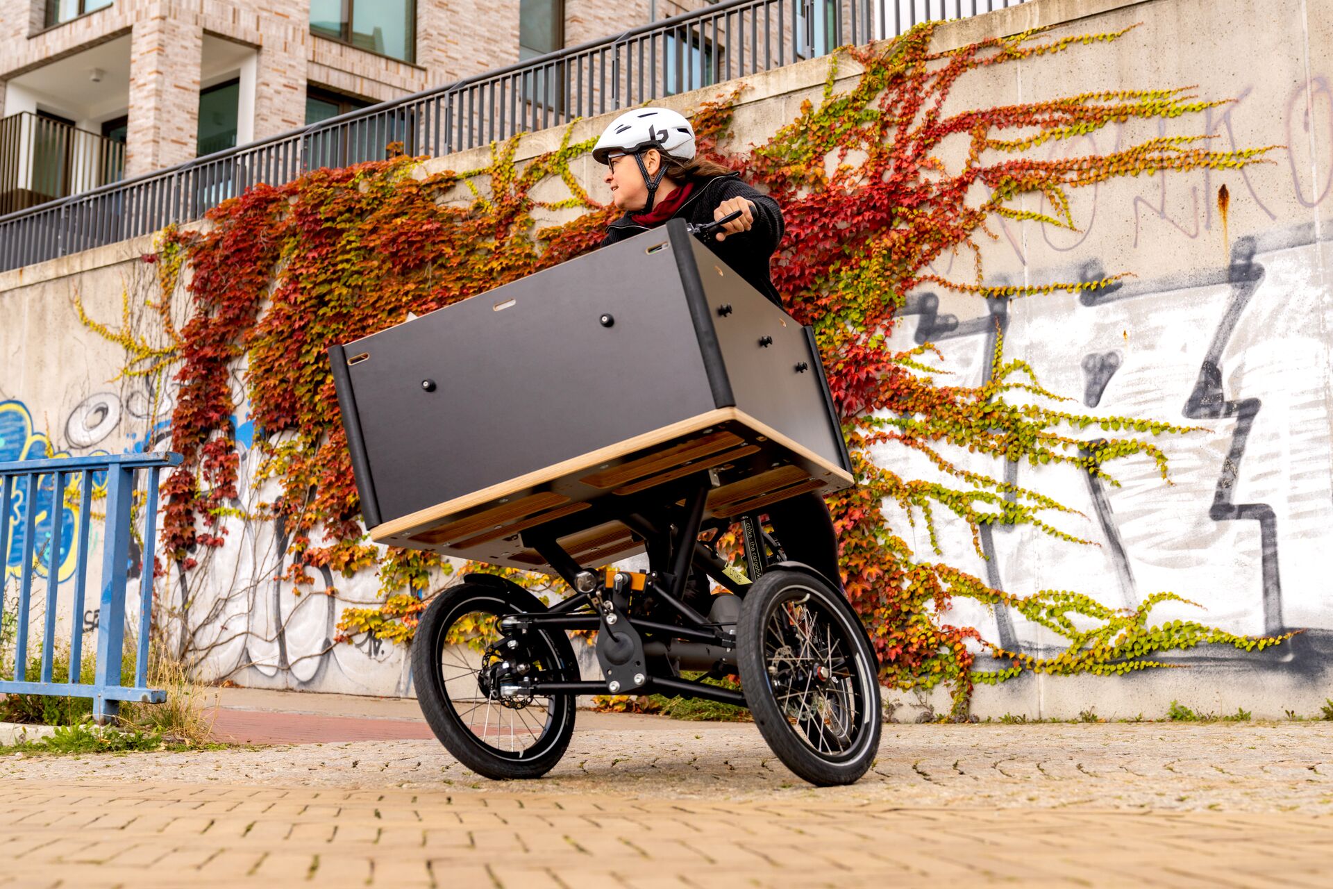 Person mit Helm fährt ein dreirädriges Lastenrad mit großer Transportbox vor einer bewachsenen Wand.